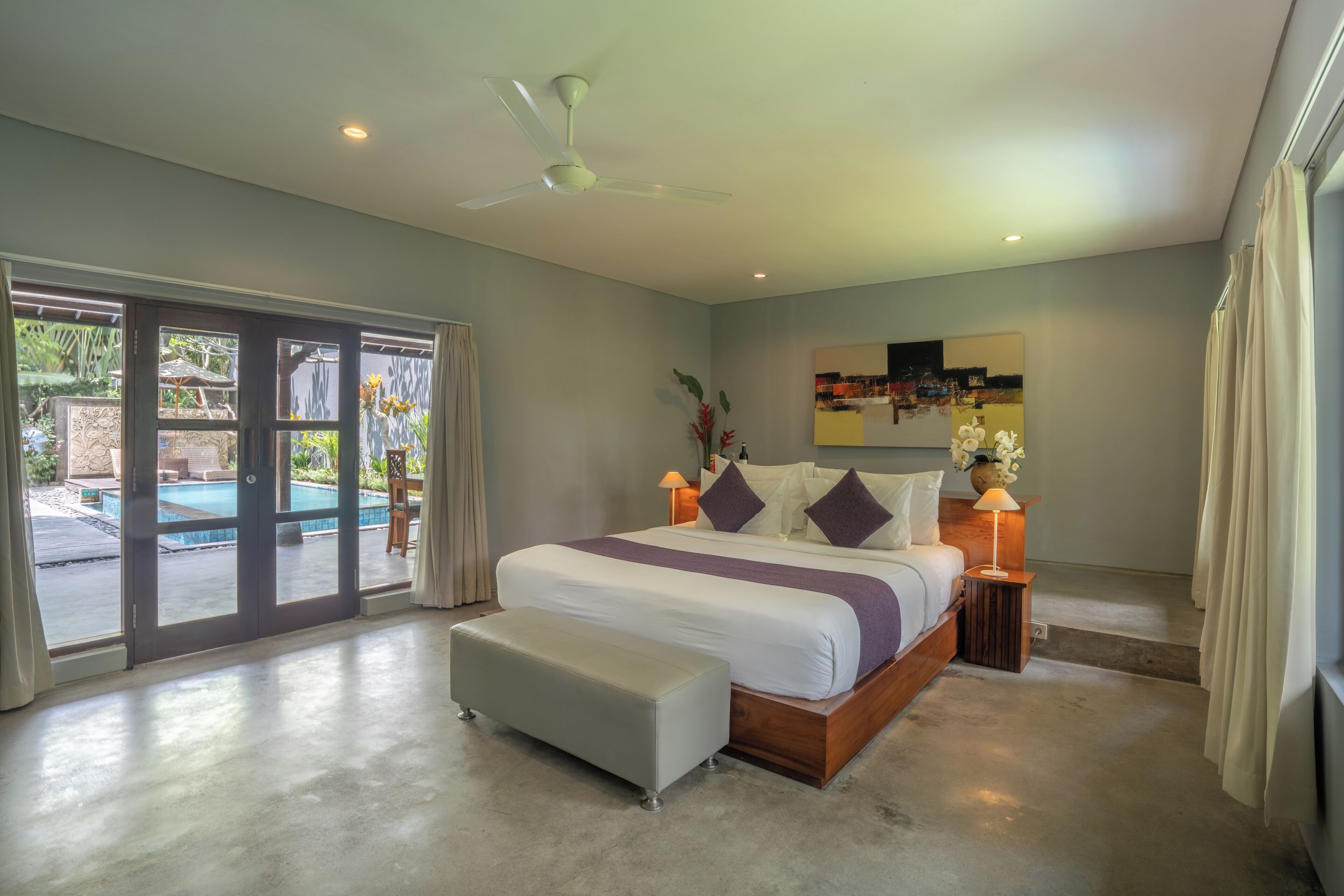 A bedroom with a large bed, a ceiling fan, a nightstand with a lamp, and a view of a swimming pool through glass doors.