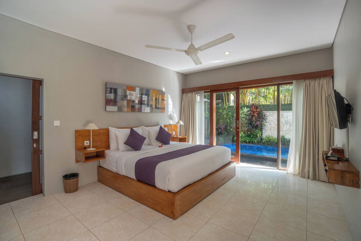 A modern hotel room with a large bed, a ceiling fan, a TV, and a sliding glass door to a garden.