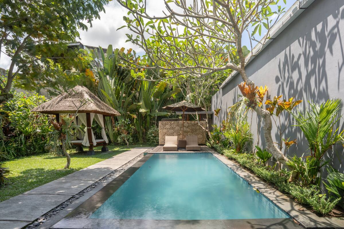 A swimming pool with a thatched gazebo, a stone wall, and lush tropical plants.