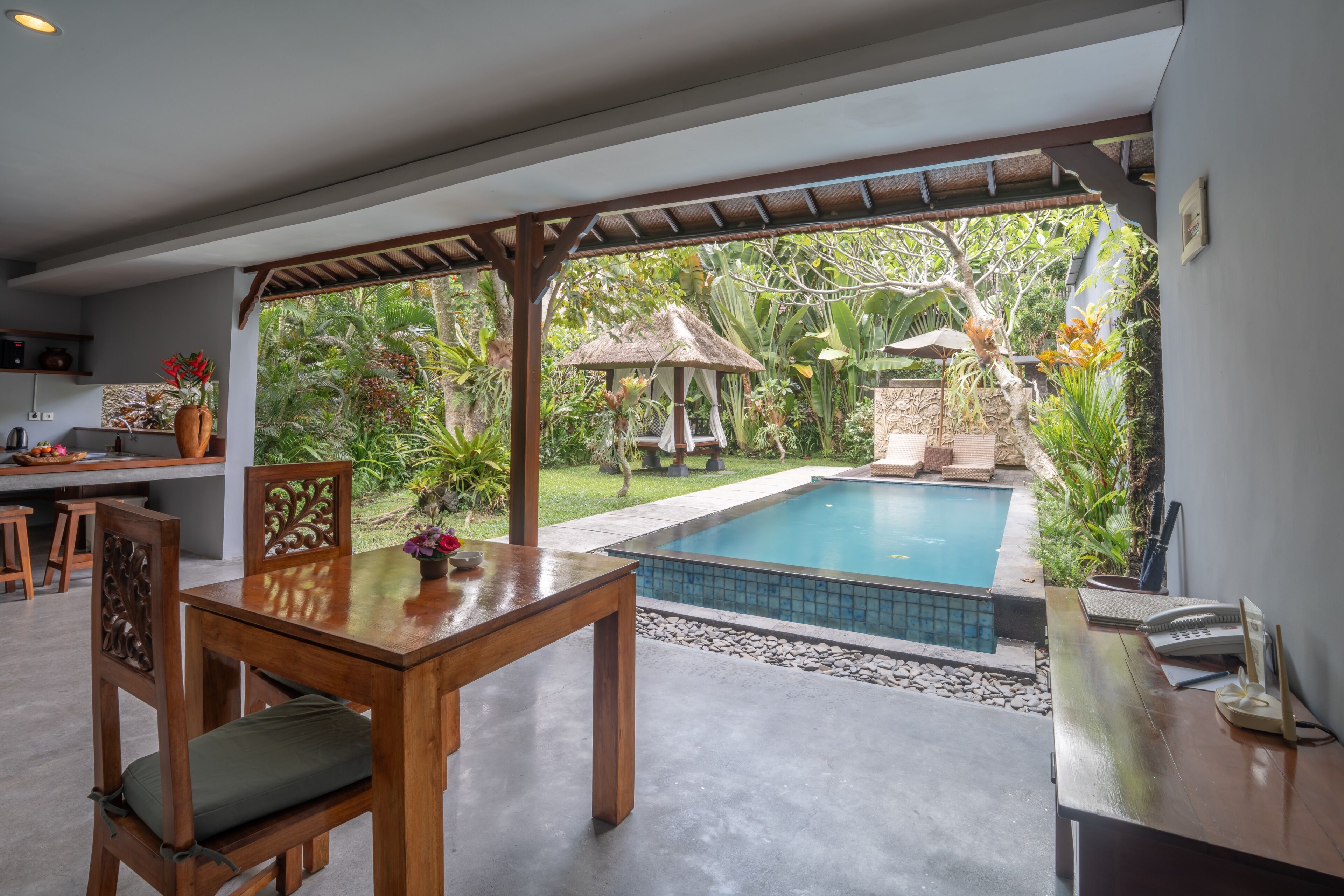 A poolside area with a wooden table and chairs, a small gazebo, and a view of lush greenery.