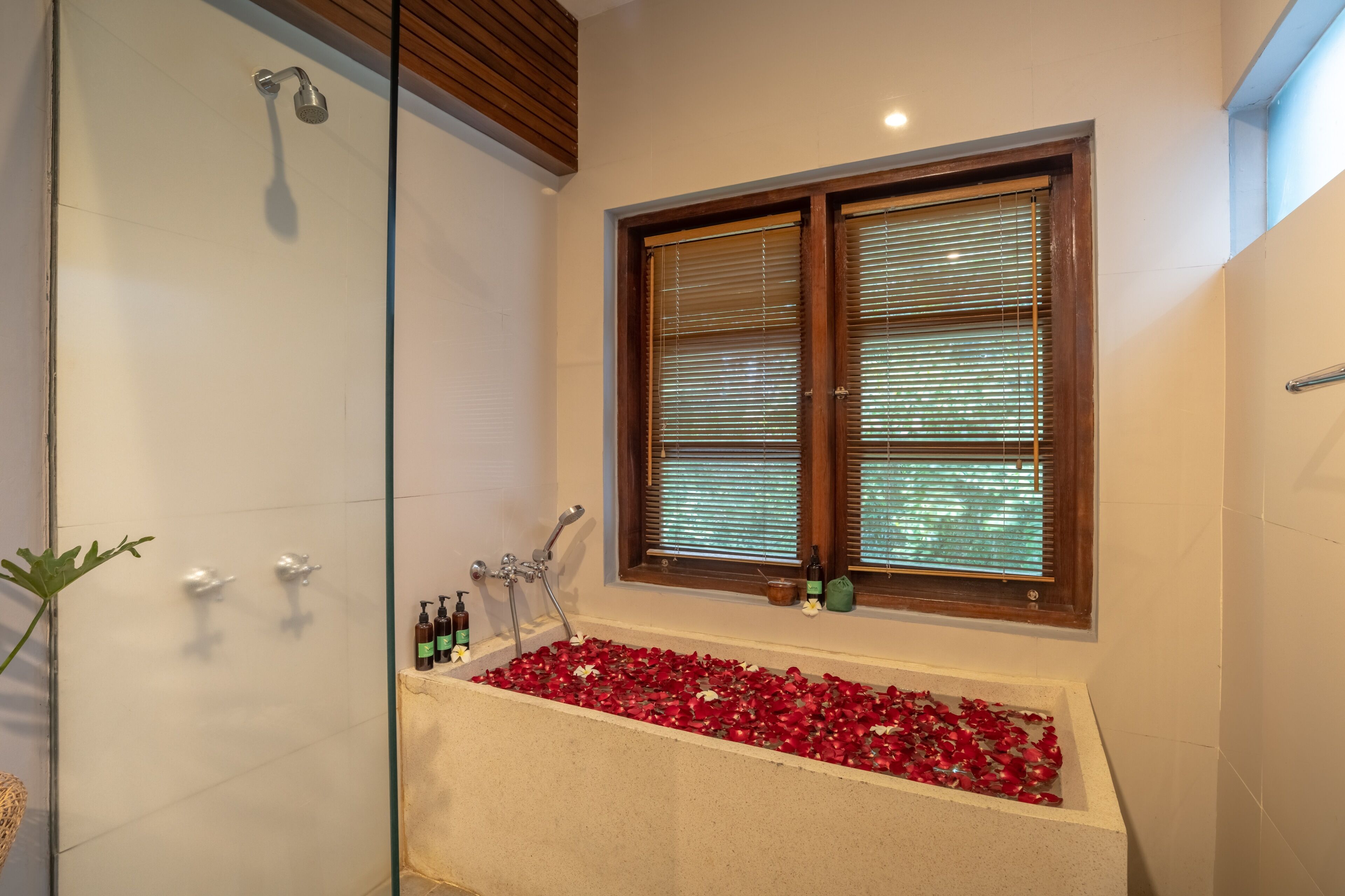 A bathroom with a glass-enclosed shower, a bathtub filled with rose petals, and a window with blinds.