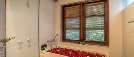A bathroom with a glass-enclosed shower, a bathtub filled with rose petals, and a window with blinds.