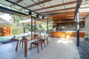 A modern kitchen with a central island, a dining area with a table and chairs, and a view of a swimming pool. - Sativa Luxury Villas Ubud (Ubud)