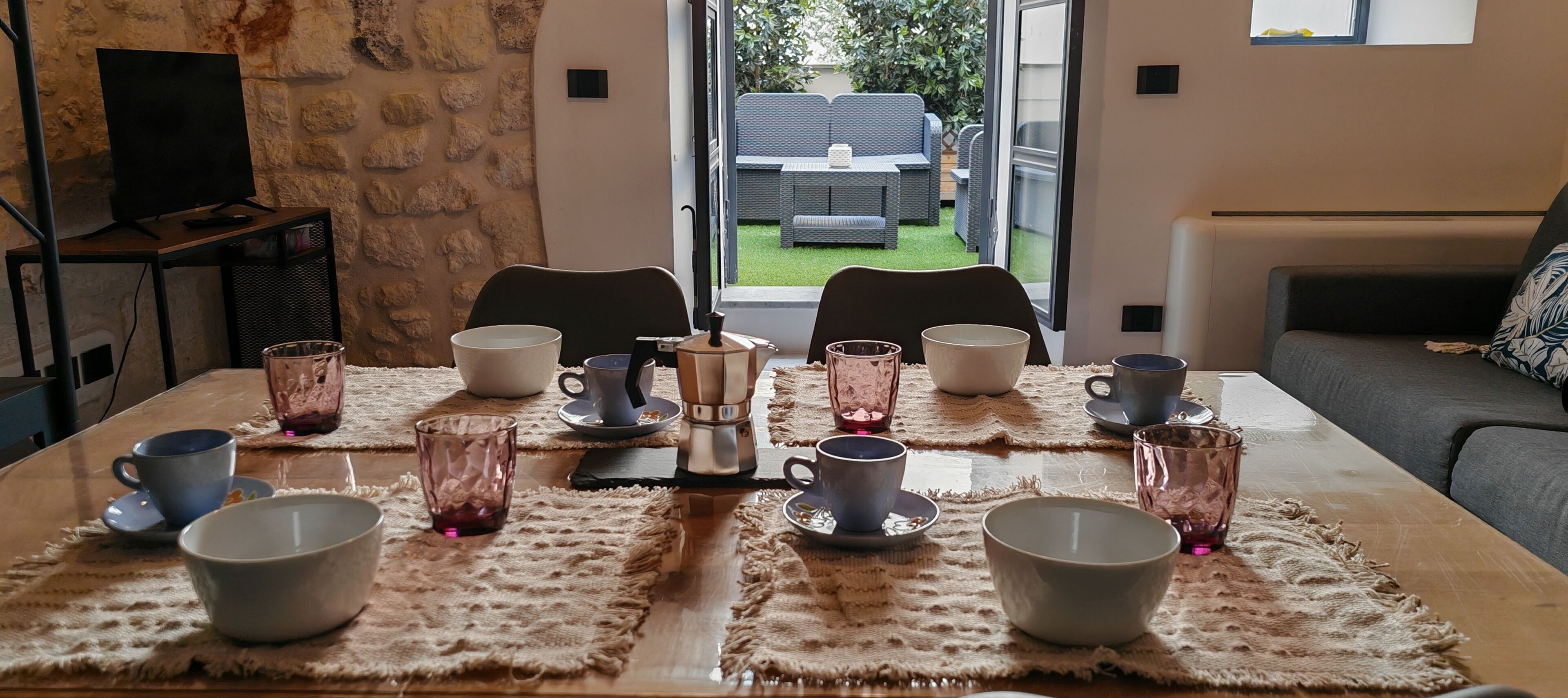 A dining table with multiple cups and saucers, a coffee pot, and a television in the background.
