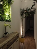 Un pasillo con piso de madera, pared blanca, planta en maceta, una silla y una mesita con un florero.