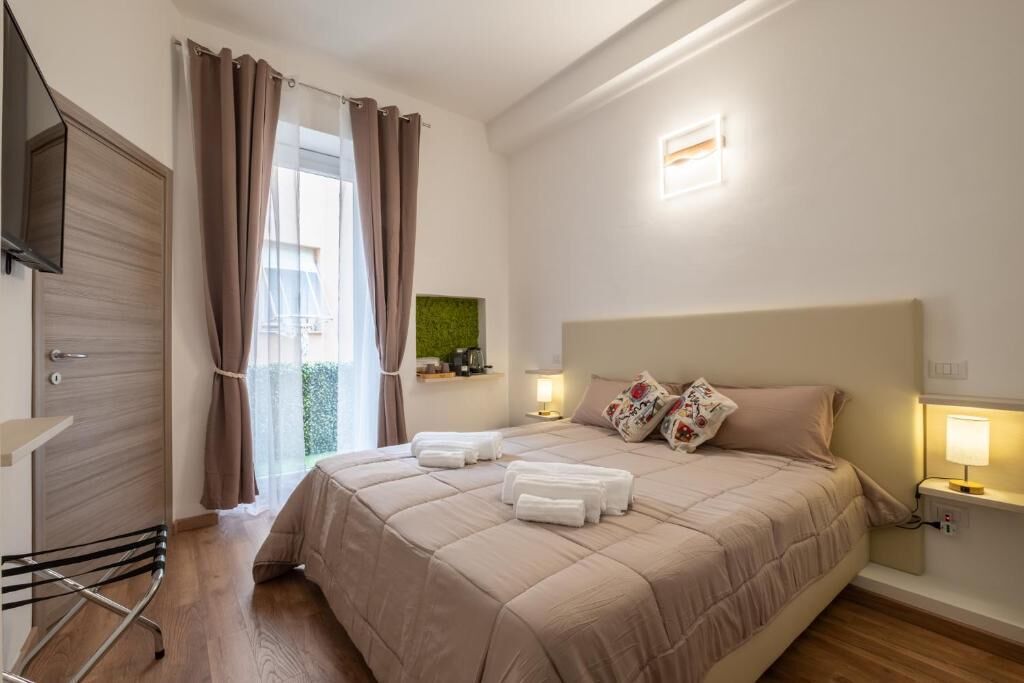Una habitación de hotel con una cama, televisión, ventana con cortinas, mesita de noche con lámpara y un perchero para toallas.