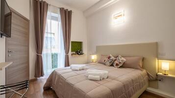 Una habitación de hotel con una cama, televisión, ventana con cortinas, mesita de noche con lámpara y un perchero para toallas.