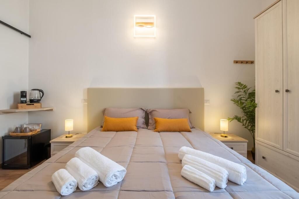 Ein Schlafzimmer mit einem Bett, zwei Kissen, einem Nachttisch mit Lampe, einem kleinen Schrank und einer Pflanze.