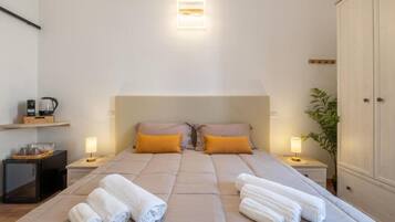 Ein Schlafzimmer mit einem Bett, zwei Kissen, einem Nachttisch mit Lampe, einem kleinen Schrank und einer Pflanze.