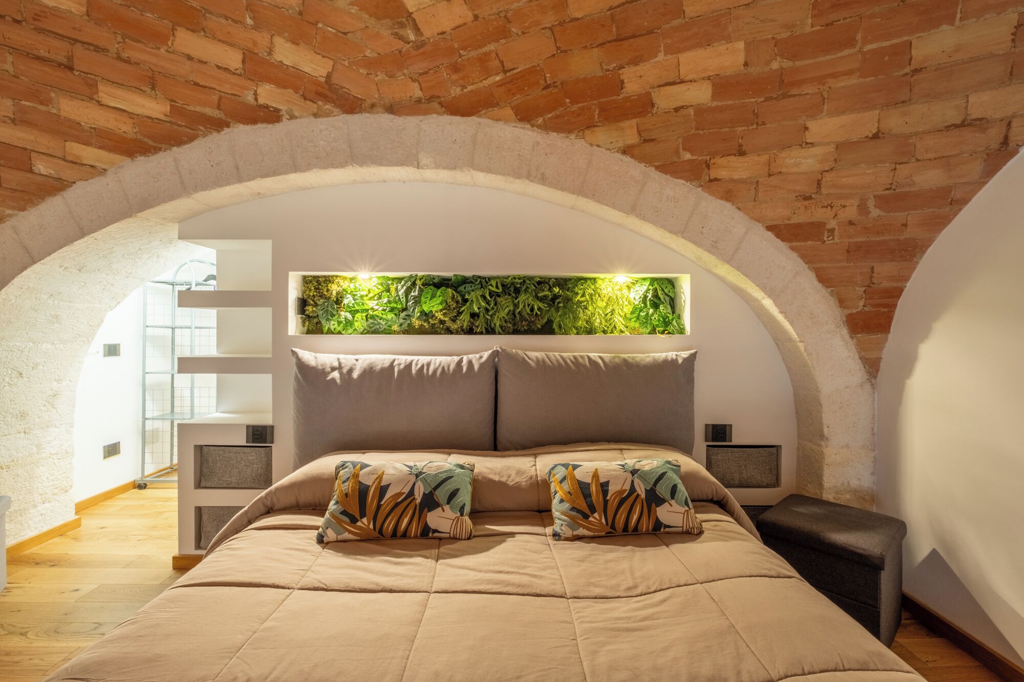 A bedroom with a brick ceiling, an arched window, a bed with a beige bedspread, and a green wall feature.