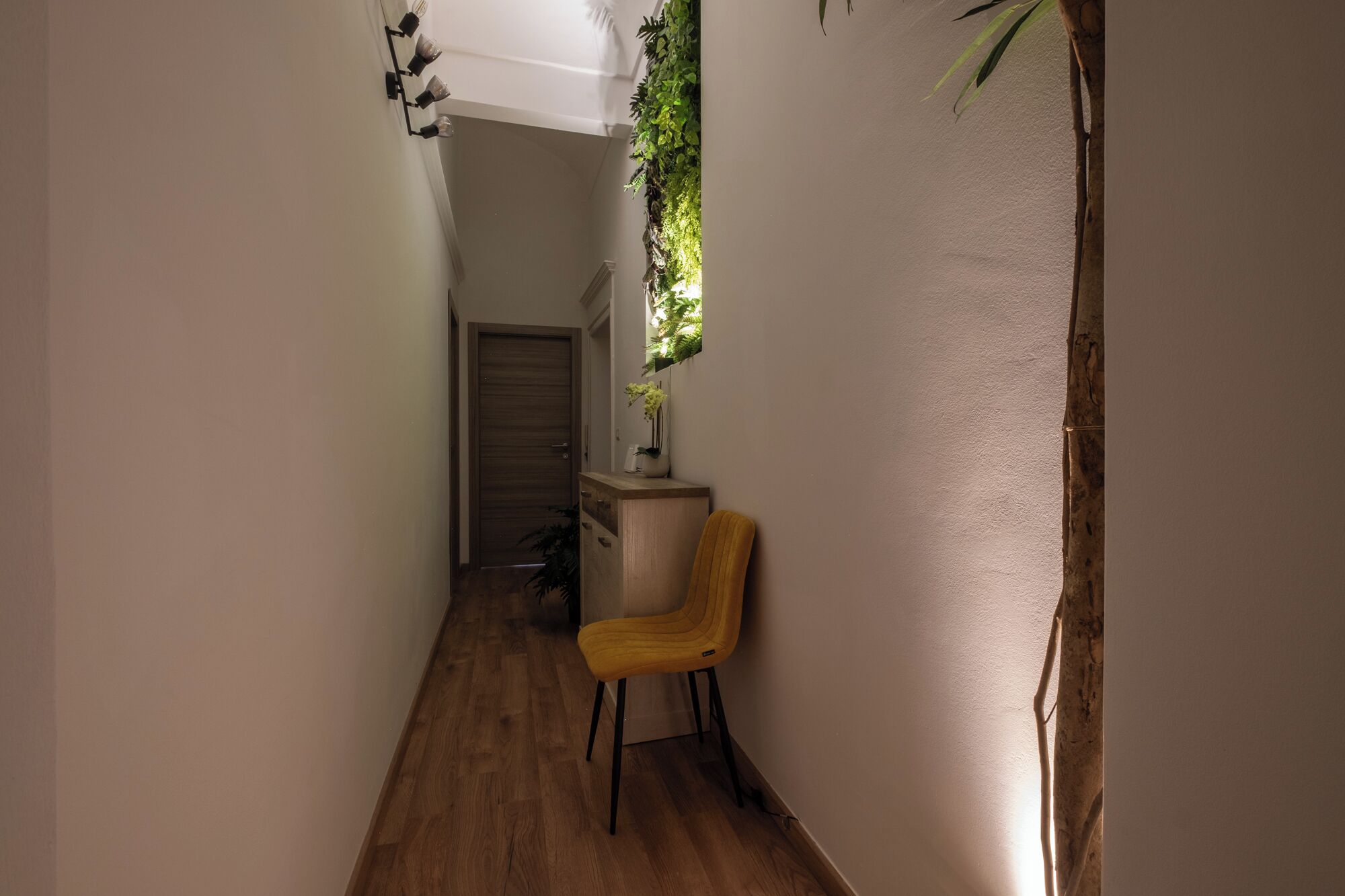 A narrow hallway with a wooden floor, a yellow chair, a small table with a vase, and a wall-mounted plant.