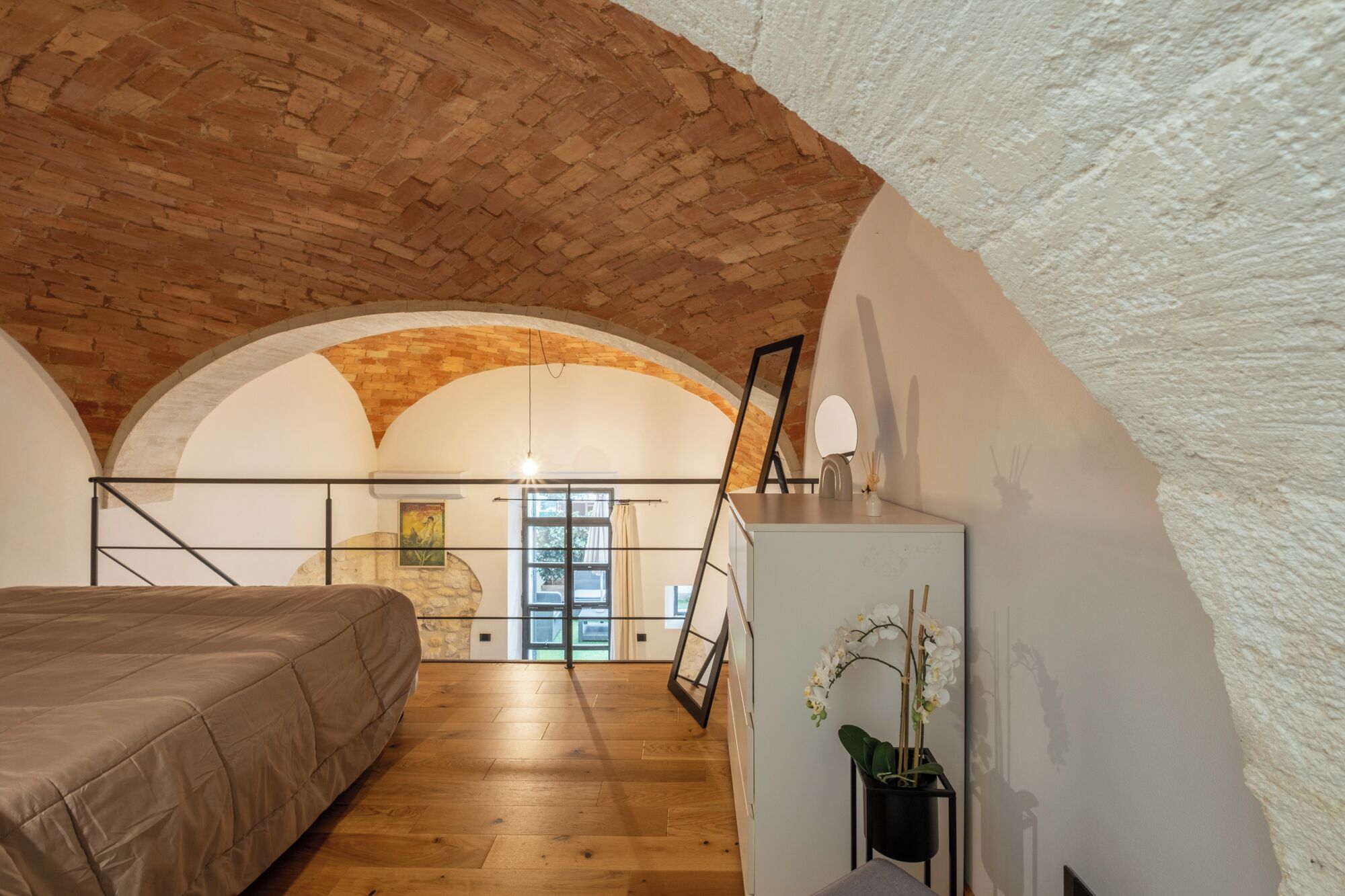 A bedroom with a wooden floor, a bed, a desk with a lamp, and a view through an arched window.