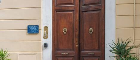 Dos puertas de madera dobles con un arco de metal decorativo encima, una placa azul en la pared y dos plantas en macetas a cada lado.