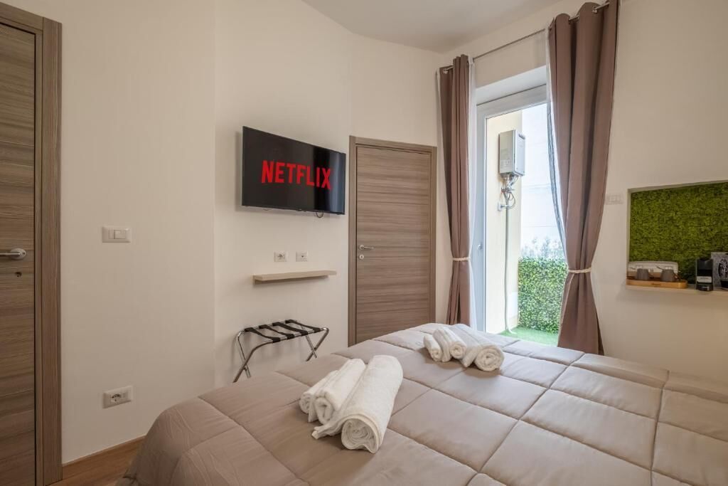 Habitación de hotel con cama, televisión montada en la pared, ventana con cortinas y un pequeño estante con artículos de aseo.