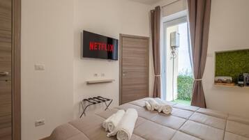 Habitación de hotel con cama, televisión montada en la pared, ventana con cortinas y un pequeño estante con artículos de aseo.