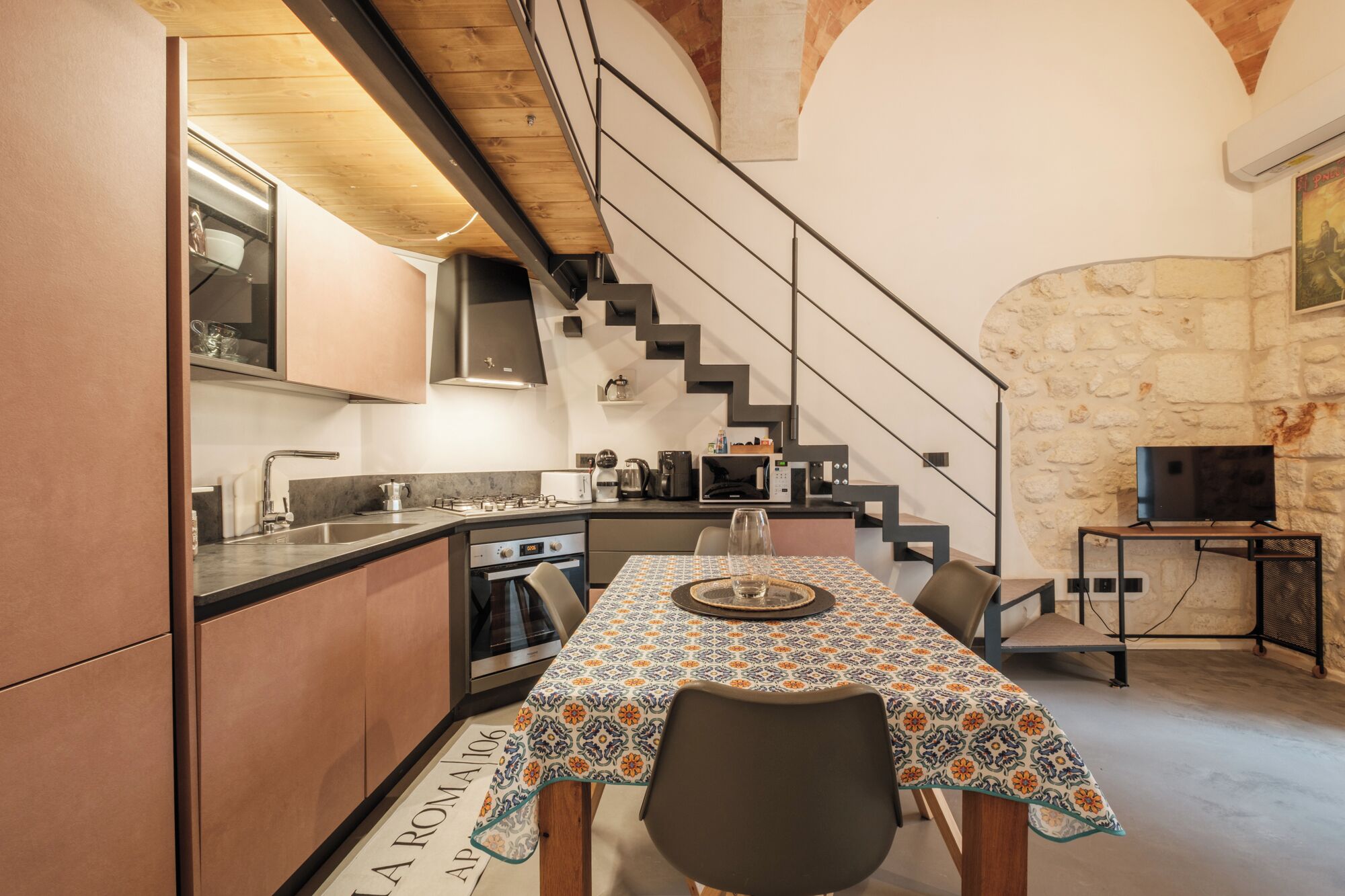A modern kitchen with a dining area, a staircase, and a stone wall.