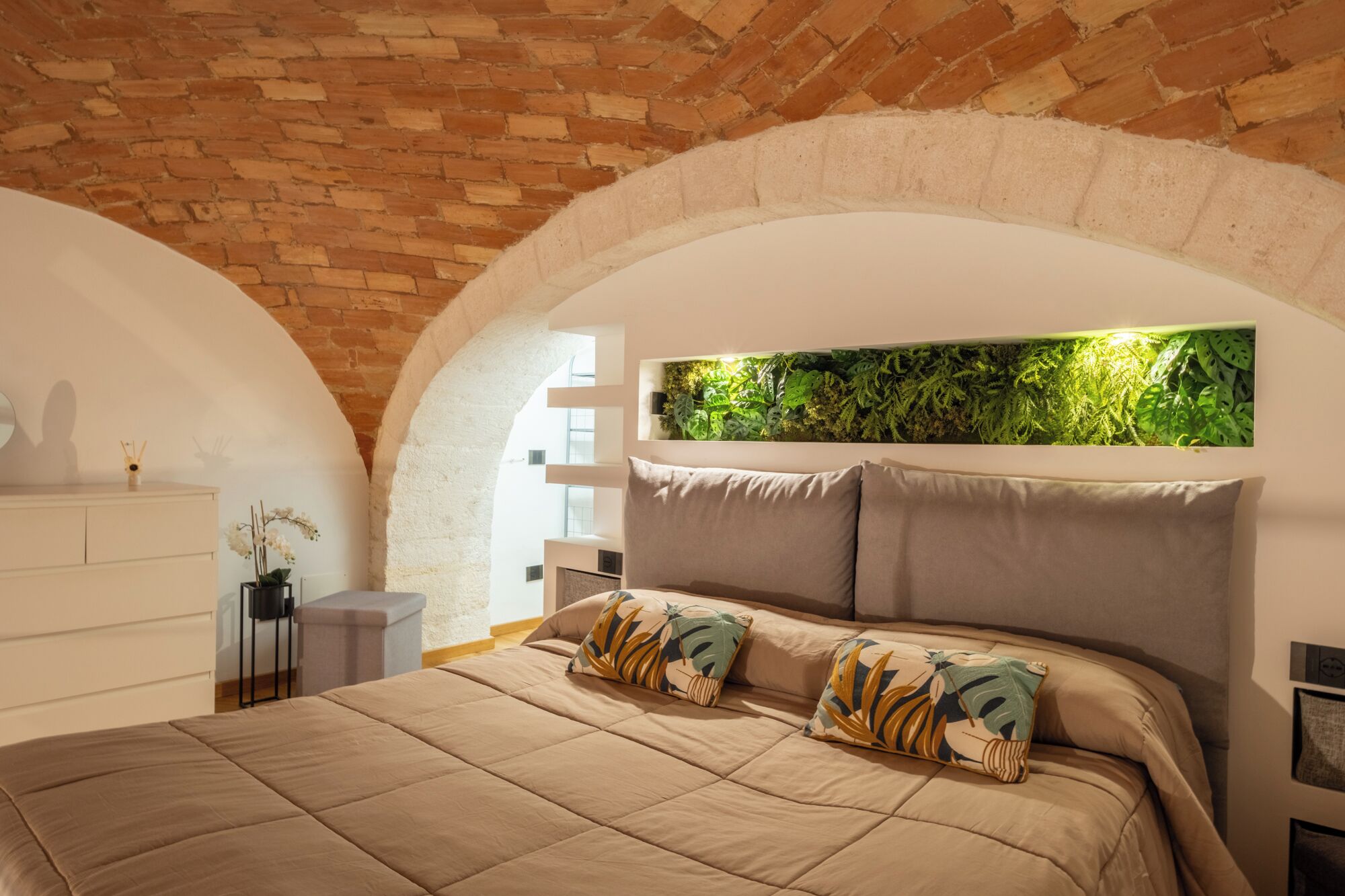 Een slaapkamer met een bakstenen plafond, een bed met een beige sprei en een plant aan de groene wand.