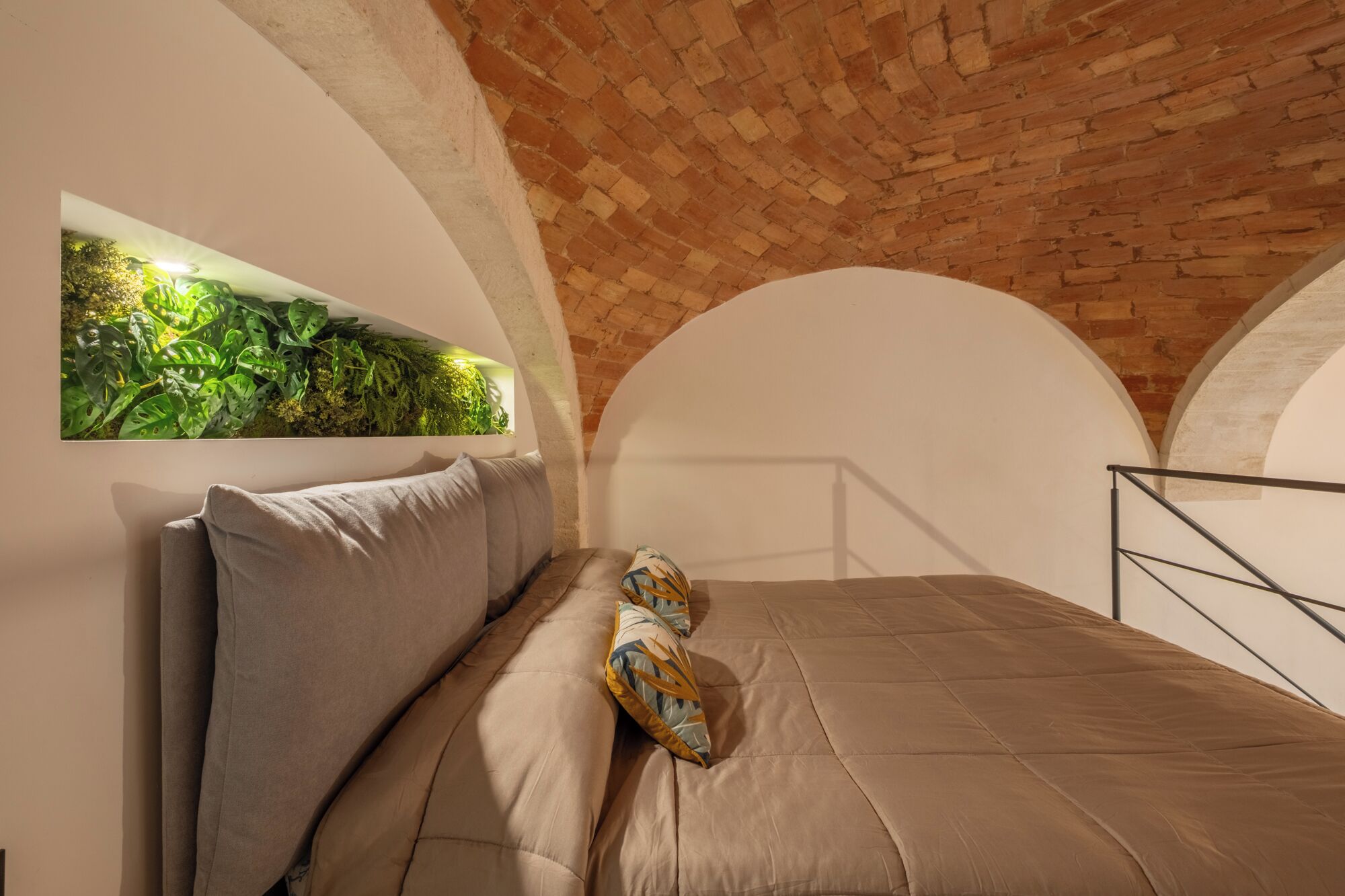Camera da letto con soffitto in mattoni, un letto con copripiumino marrone, una pianta fissata al muro e una ringhiera in metallo.