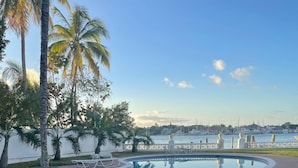 Una piscina con un sendero de piedra que la atraviesa, rodeada de palmeras y un puerto deportivo al fondo.