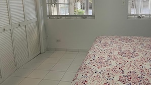 Un dormitorio con una cama, dos ventanas con persianas, un aire acondicionado blanco y suelo de baldosas.