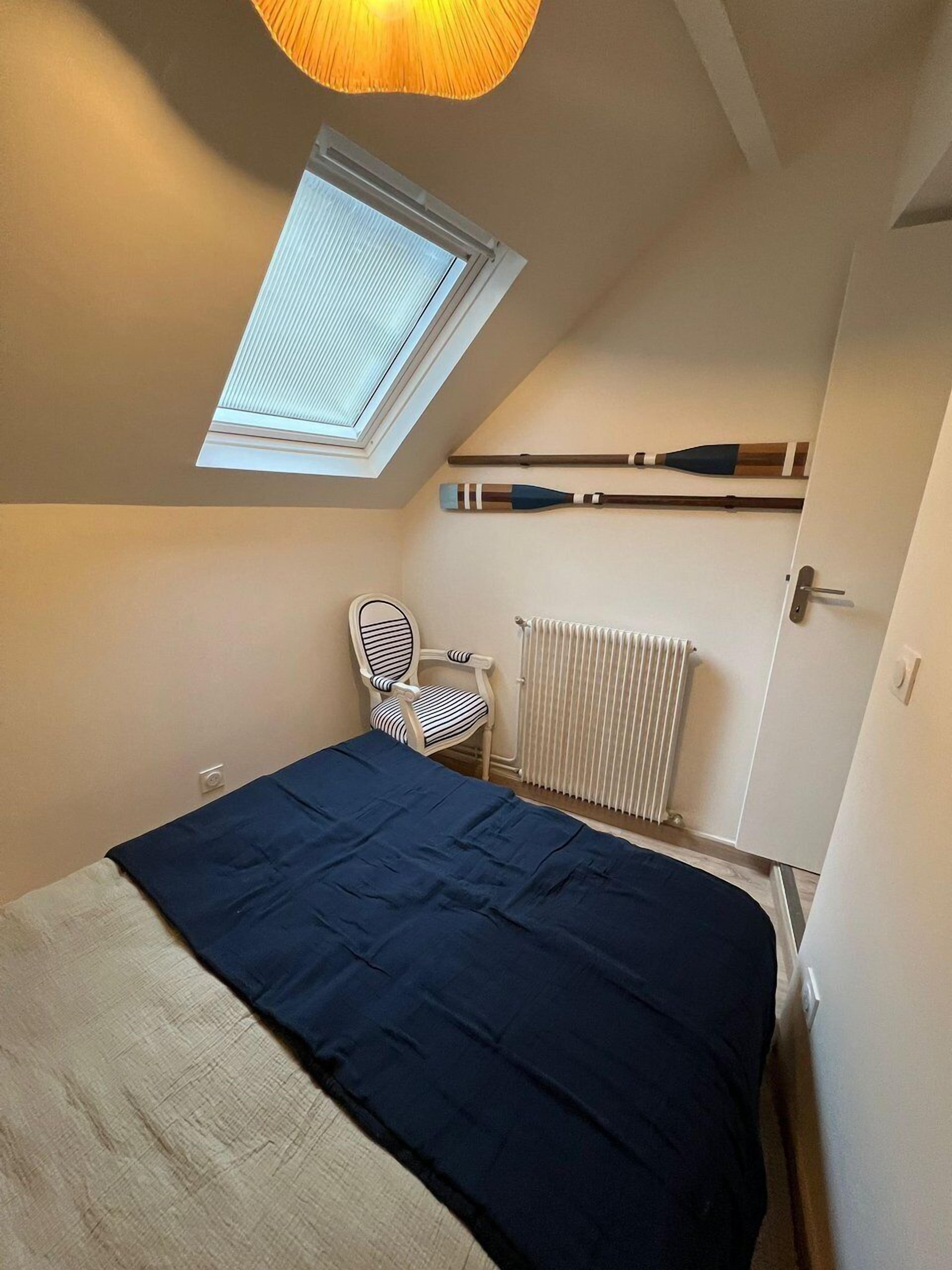 Une petite pièce agréable, avec un plafond en pente, un puits de lumière, un lit recouvert d’une couverture bleue, une chaise à rayures et un radiateur blanc.