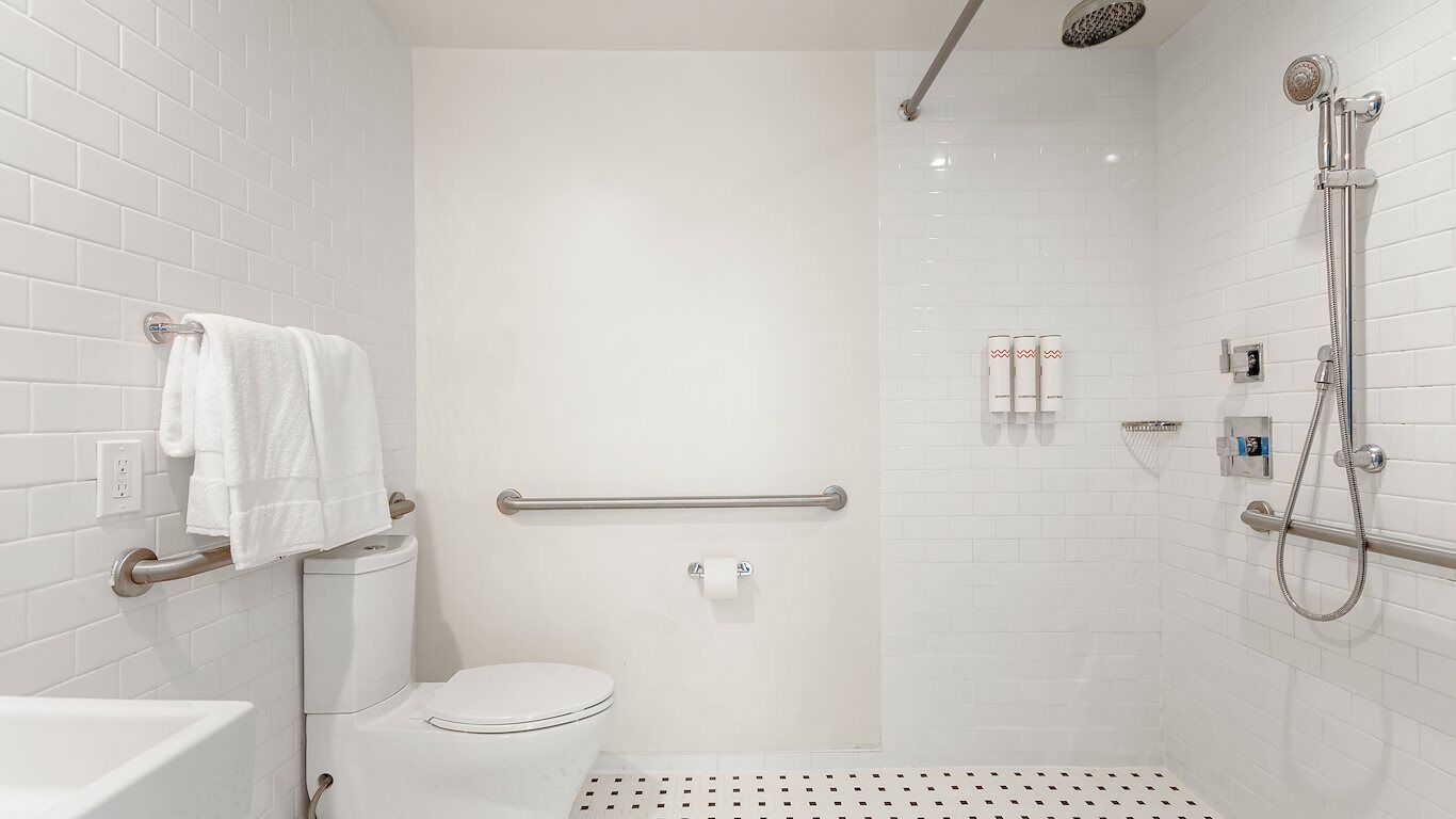 A modern bathroom with white tiles, a toilet, a shower with a handheld showerhead, and a grab bar.