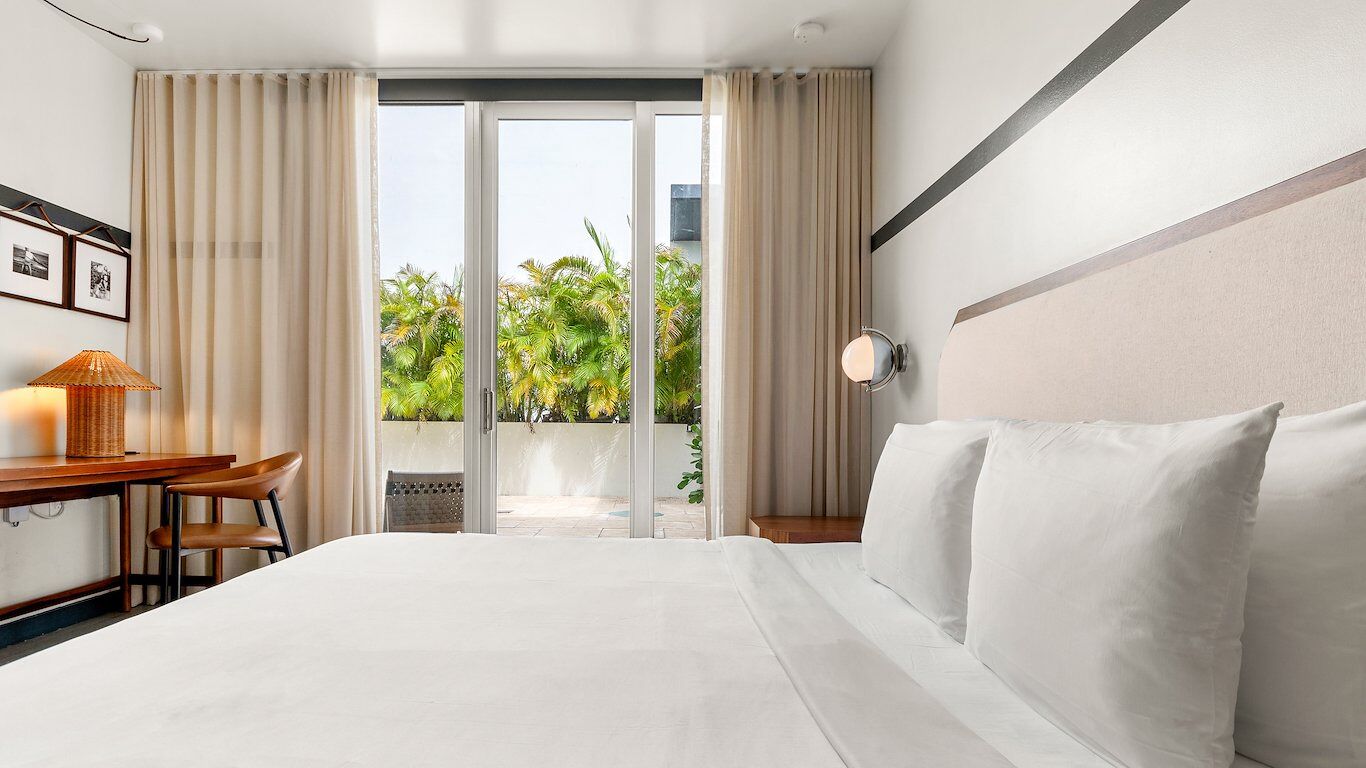 A hotel room with a bed, a desk with a lamp, a chair, and a view of greenery through a sliding door.