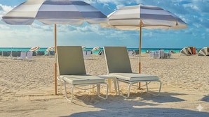 Twee strandstoelen met parasols, een zandstrand en tenten op de achtergrond.