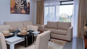 Ein stilvolles Wohnzimmer mit einer Couch, einem Esstisch und Stühlen. Es gibt ein großes Fenster mit Vorhängen, ein Gemälde an der Wand und einen Blick auf Gebäude im Freien.
