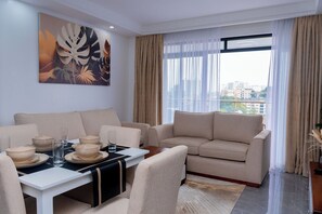 Een moderne woonkamer met een bank, een eettafel en stoelen. Er is een groot raam met gordijnen, een schilderij aan de muur en een uitzicht op gebouwen.