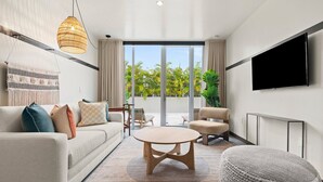 Een moderne woonkamer met een bank, salontafel, stoelen, een aan de muur bevestigde televisie en een groot raam met uitzicht op groen.