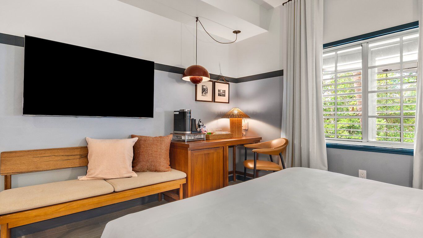 Une chambre d’hôtel avec un lit, un bureau avec une lampe, un canapé, une télévision et une fenêtre donnant sur un paysage verdoyant.