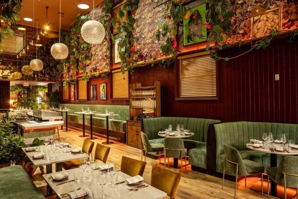 A restaurant with green upholstered booths, wooden tables, hanging spherical light fixtures, and a wall adorned with plants and artwork.