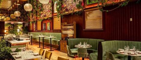 A restaurant with green upholstered booths, wooden tables, hanging spherical light fixtures, and a wall adorned with plants and artwork.