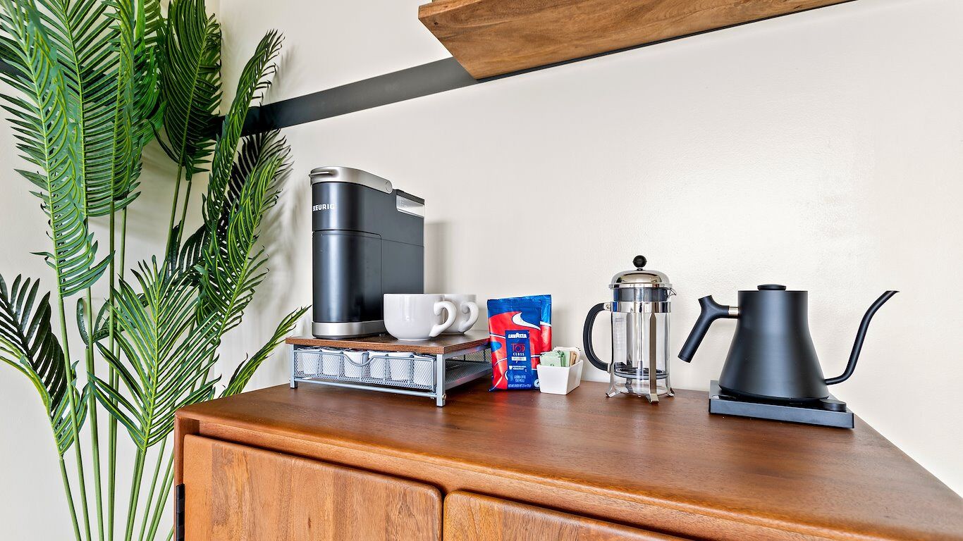 A coffee maker, a French press, a cup, a box of coffee pods, and a kettle on a wooden surface.