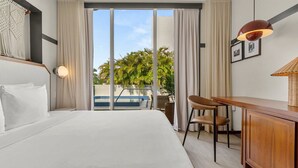 A hotel room with a bed, a desk, a chair, a lamp, and a view of a pool and palm trees. - The Daydrift | 2BR Suite | Close to the Pulse, Comfortably Apart | 2 Units (Miami Beach)