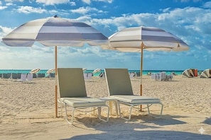 Two beach chairs with umbrellas, a sandy beach, and tents in the background. - King Terrace | The Daydrift | Rooftop Pool, Lounge Deck, Collins Park (Miami Beach)