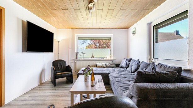 A modern living room with a wooden ceiling, a large window with a view, a flat-screen TV, a sofa, and a coffee table with a bottle and glasses.