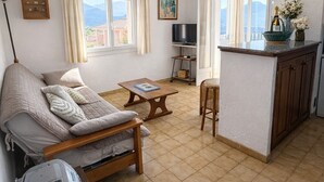 A cozy living room with a sofa, a coffee table, a TV, and a kitchen area with a counter and a window. - PROMO - Sea & mountain view - 6p - parking - Linen included (Corbara)