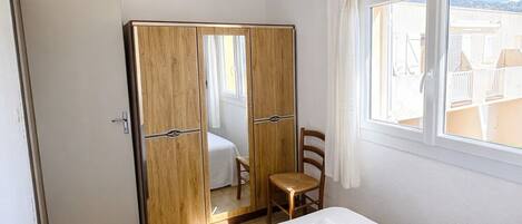A hotel room with a bed, a wooden wardrobe, a small table with a chair, and a window with curtains.