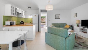 Een moderne keuken met witte kasten, een magnetron en een koffieapparaat. Er is een eethoek met een witte tafel en stoelen. Een groene bank staat in de woonkamer, en er is een televisie op een witte dressoir.