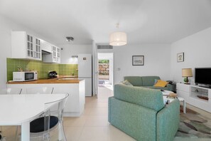 Une cuisine moderne avec des meubles blancs, un four à micro-ondes et une machine à café. On y trouve un coin repas avec une table et des chaises blanches. Un canapé vert se trouve dans le salon, et un téléviseur est posé sur un meuble blanc.