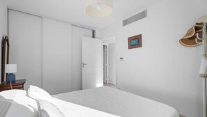Een slaapkamer met een groot bed, een nachtkastje met een lamp, een deur naar een andere kamer en een hoedenrek.