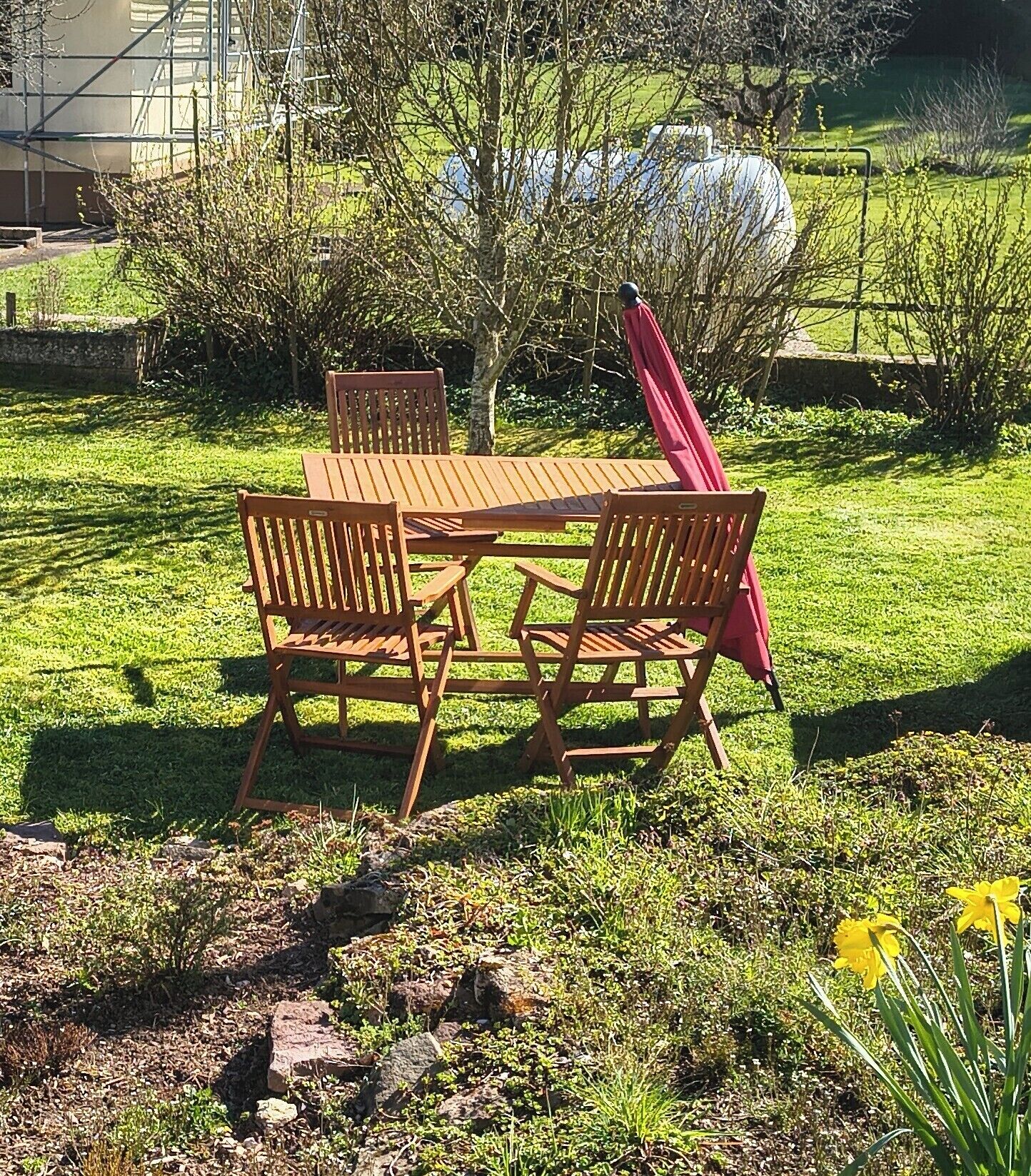 Un espacio al aire libre con mesa y sillas de madera, rodeado de follaje y un muro de piedra.