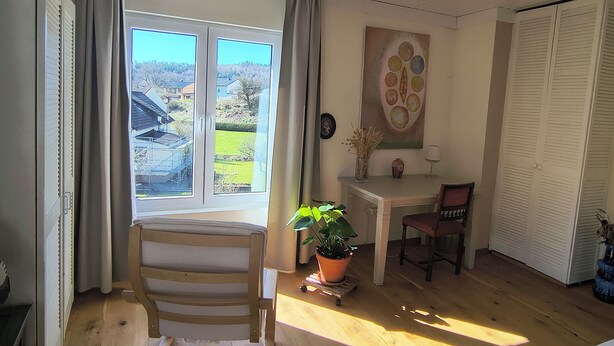 Ein Zimmer mit Holzboden, einem fenster mit Vorhängen, einem Stuhl, einem Tisch mit Vase und Lampe sowie einer Tür mit Lamellen.