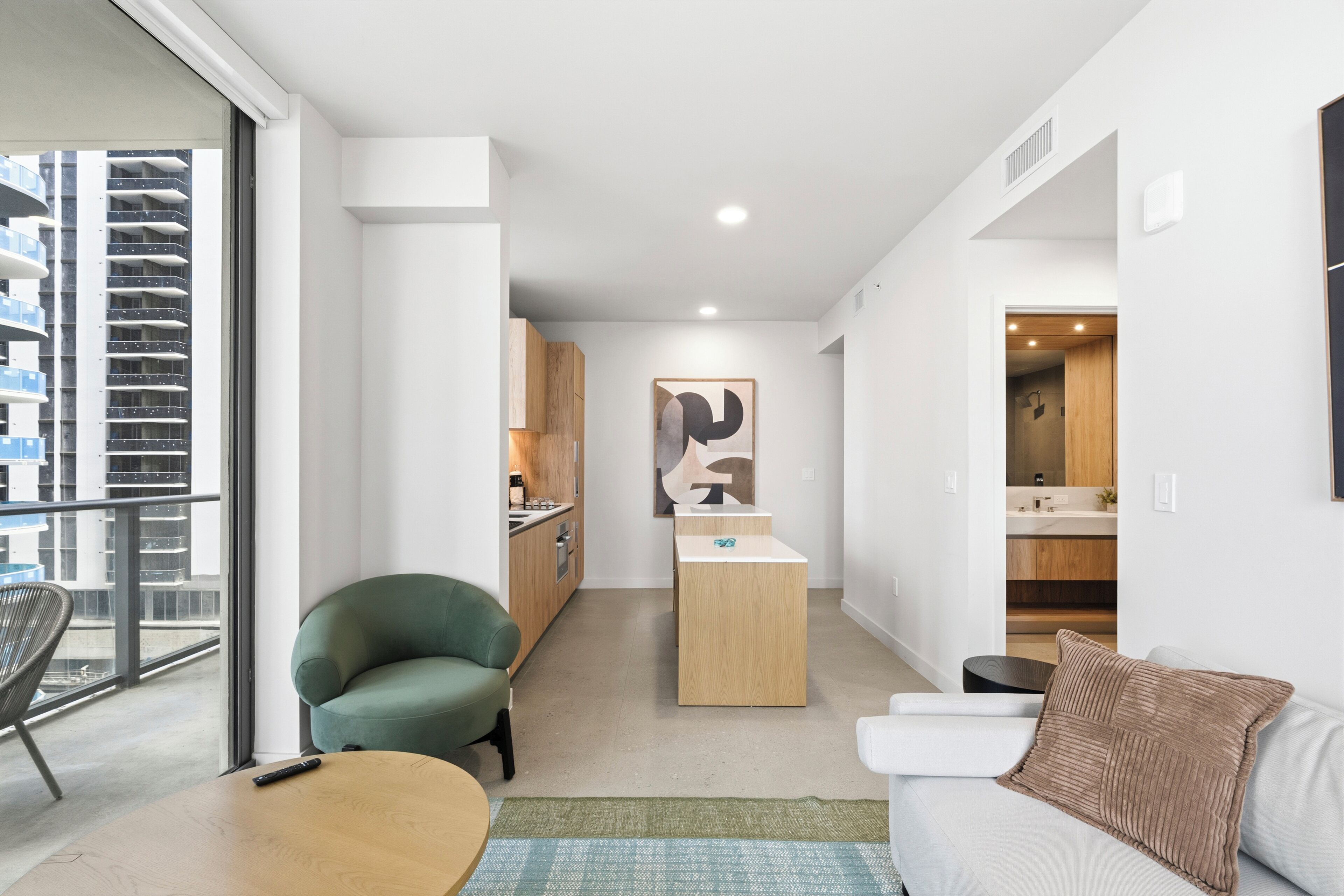 A modern apartment with a balcony, a green armchair, a wooden coffee table, a sofa with a brown cushion, and a kitchen area in the background.