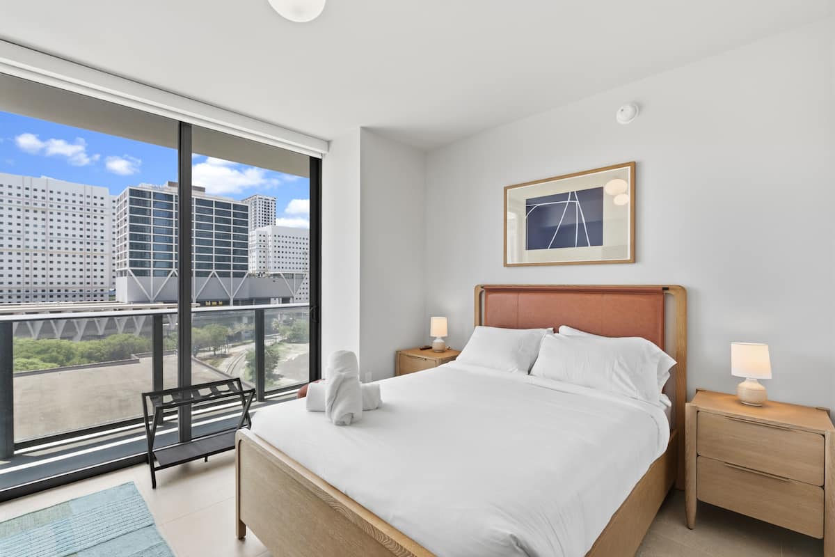 Une chambre d’hôtel moderne avec une grande fenêtre, un lit recouvert de draps blancs, une tête de lit en bois, une table de chevet avec une lampe et un tableau encadré au mur.