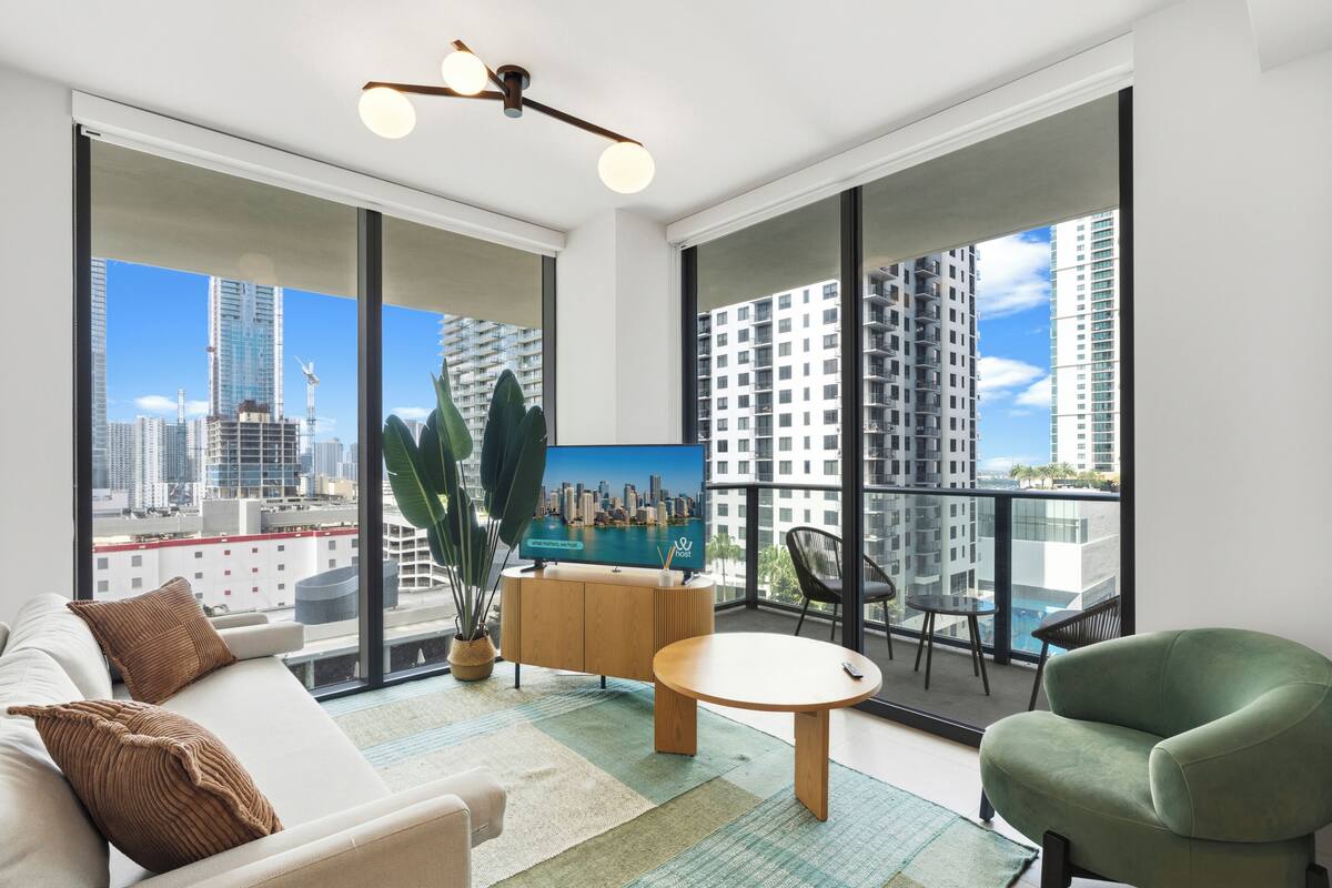 Un salon moderne avec un canapé, une table ronde, un fauteuil vert, un téléviseur et un balcon offrant une vue sur la ville.