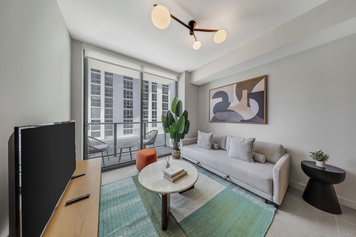 Un salon moderne avec un téléviseur à écran plat, une table basse ronde, un canapé, une table d’appoint avec une plante et un grand tableau abstrait accroché au mur.