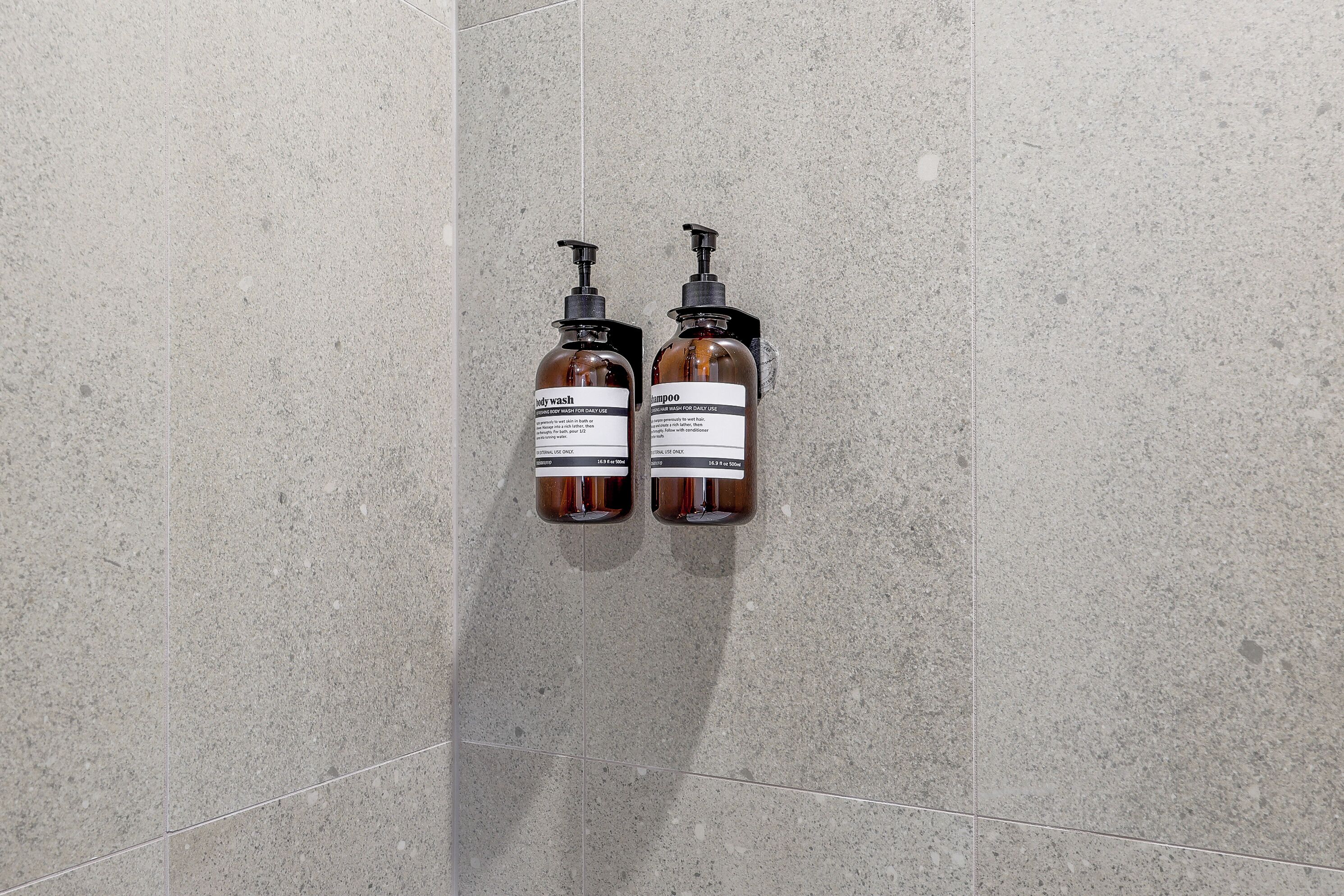 Two bottles of body wash and shampoo mounted on a tiled wall.